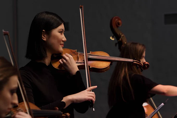 A photo of female violinist performing on stage during a live concert. There are two other musicians on either side of her playing a violin and a double bass.