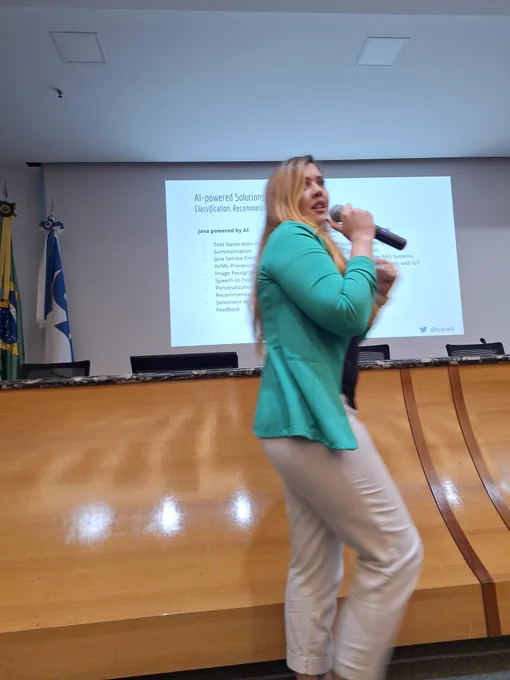 mulher loira de calça clara e blazer verde fala ao microfone enquanto se movimenta em frente a um auditorio