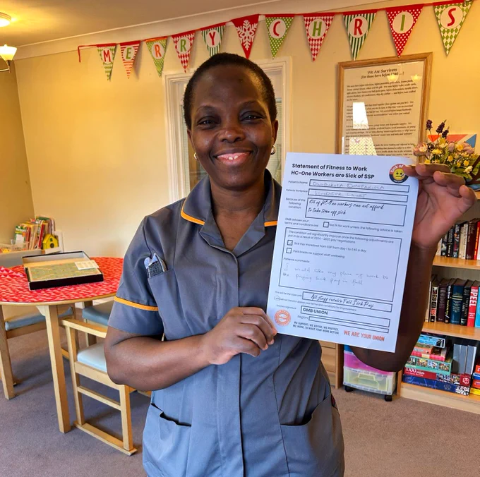 A smiling care worker in a home holds up a sick note campaign prop calling for improved sick pay and paid breaks for care workers.