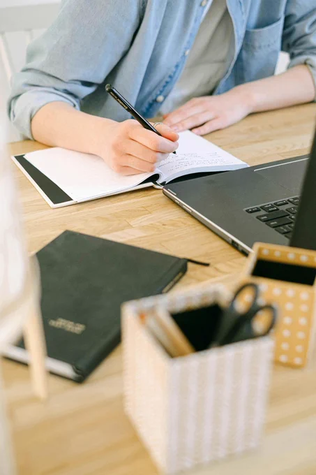 A far distant of a person sitting at a desk writing in a notebook with a laptop in front of them.