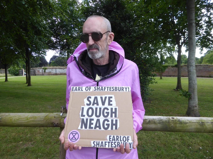 A man wearing a pink hazmat suit  (one of XR Wimborne’s Pink Rebels) holds a placard which reads “Earl of Shaftesbury, Save Lough Neagh”. The photograph was taken outside St Giles House in Wimborne St Giles, the home of the Realisation Festival