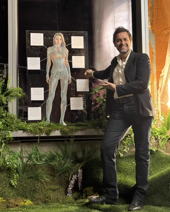 A designer stands next to a poster with a drawing of a concept. He is gesturing toward the poster and standing on a foreground of faux greenery and ferns.