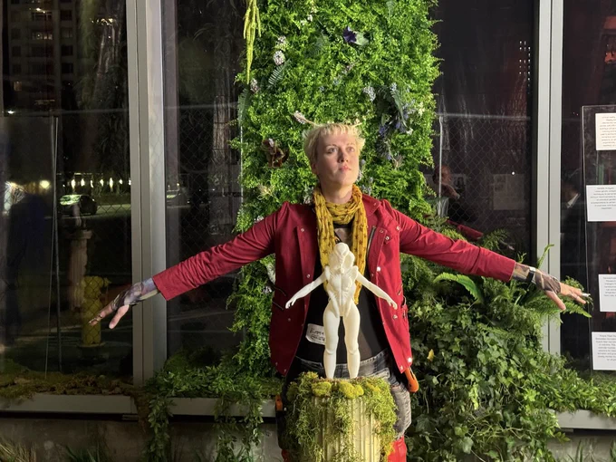 A team member poses with arms outstretched and face to the sky, mimicking the pose of statuette designed to model the garment concept. Faux foliage decorates the background. 