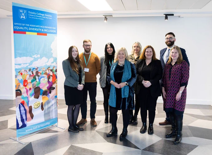 Group photo of Trinity's Equality, Diversity and Inclusion Team, standing beside a pull-up banner with a colourful painting of a crowd of people. Text on the banner reads 'Office of the Associate Vice-Provost for Equality, Diversity & Inclusion', plus the Trinity logo.