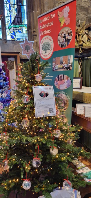 Christmas tree decorated with lights, photo baubles and a star with EMAST's logo (the words 'East Midlands Asbestos Support Team' around a tree). Behind the tree is a banner featuring the words 'Justice for Asbestos Victims', the EMAST logo and photos of staff and service users.