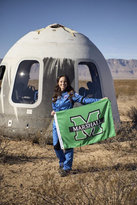 Emily holding a Marshall University flag in front of a landed Blue Origin capsule