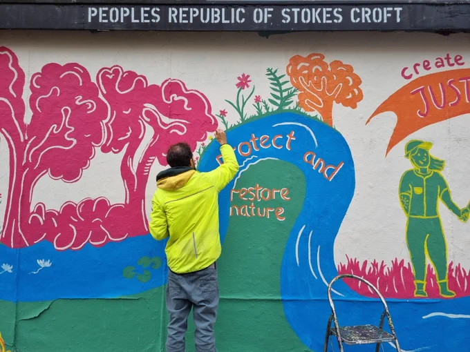 A close up of the mural being painted. "Protect and restore nature", with a background of a river, trees and plants