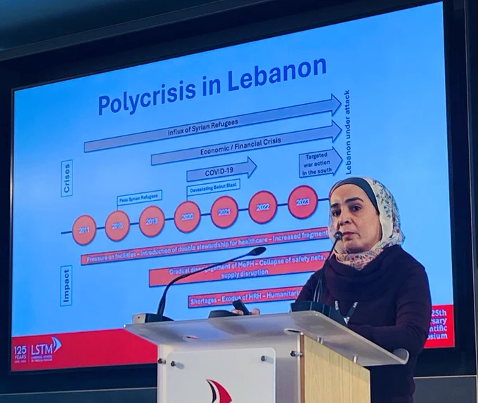 A woman stands speaking at a lectern with a screen behind showing the polycrisis in Lebanon