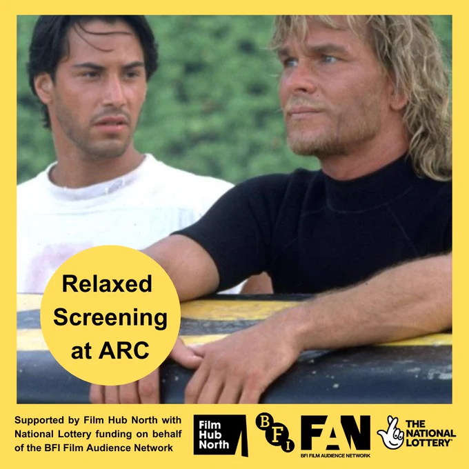 Still from Point Break. Keanu Reeves wearing a white T-shirt looks over at Patrick Swayze, who is wearing a black wetsuit and holding a surfboard.

 At the bottom are the Film Hub North, BFI, Film Audience Network and National Lottery logos. Black text on a yellow circle over the image says: Relaxed Screening at ARC.