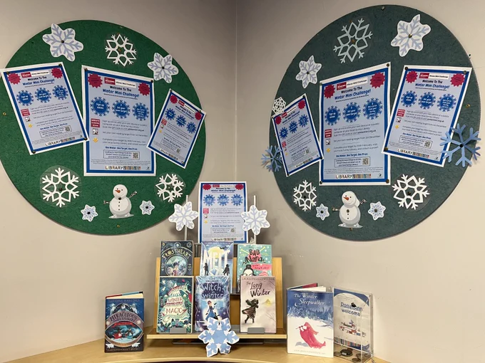 An image of two circular display boards in a Children's library. On the display boards are posters advertising the Winter Mini Challenge with snowflakes around them. Below the display boards is a book display of children fiction books with the theme of winter. 