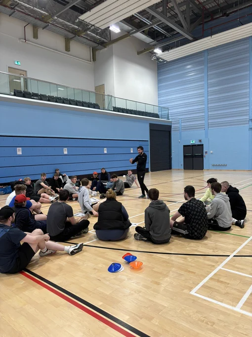Group of students sat in a sports hall being delivered information as part of a training course. 
