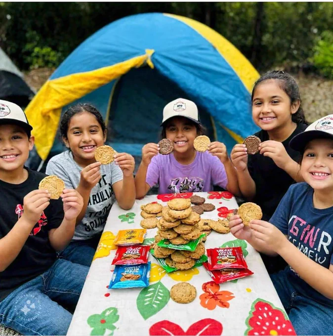 AI-generated image of Girl Scouts in front of a tent with a pile of cookies on a table.  No known children are in this AI—generated image.
