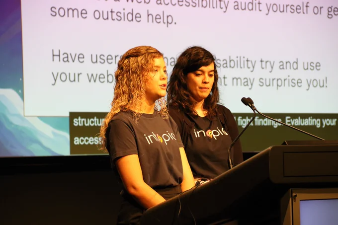 Close up of Lauren and Peyton from Intopia presenting together at the lectern. Part of their presentation is on the screen behind them.