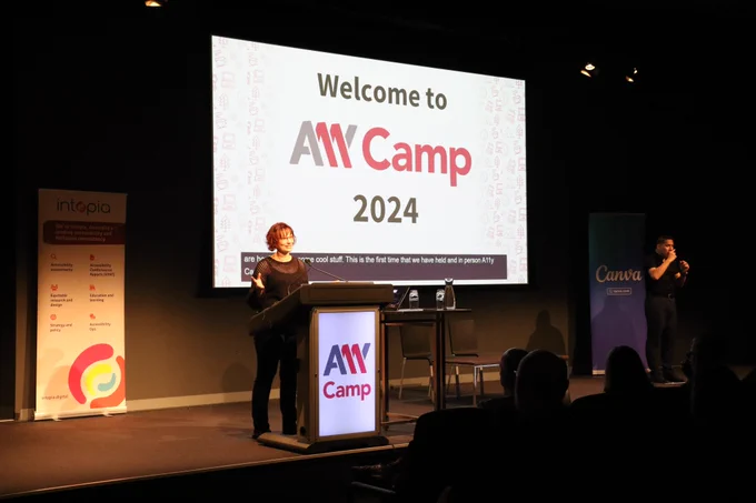 Sarah Pulis welcoming everyone to A11y Camp. She is standing at the lectern, A large screen behind her reads "Welcome to A11y Camp 2024". An Auslan interpreter is on the right of stage.