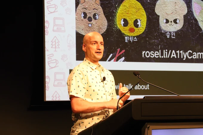 Close-up of Adrian Rosselli presenting at the lectern. Cute cartoon animal faces from his slide presentation are behind him on the big screen.