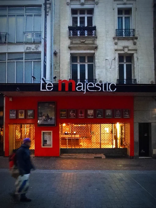 Cette photo montre la façade du cinéma Le Majestic à Lille, reconnaissable par son enseigne rouge et noire, avec le grand "m" rouge mis en avant dans le nom. On peut observer plusieurs affiches de films exposées au-dessus de l'entrée (12 petites et une grande), et à travers les grilles baissées, des escaliers illuminés menant aux étage s du bâtiment.

Au premier plan, une personne passe, légèrement floue. Elle est vêtue d'une gros veste noire et est coiffée d'un bandana violet. Elle porte un sac en plastique blanc dans la main droite et un sac à dos rouge.

L'architecture du bâtiment est élégante, avec des fenêtres traditionnelles et des balustrades, typiques des bâtiments historiques de ce type. Sur la gauche la façade est entièrement vitrée