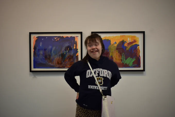 Lucy Burns smiles for the camera next to her paintings Untitled I and Untitled II, at the Sensation reception. She is wearing an Oxford University hoodie and a white crossbody bag. 