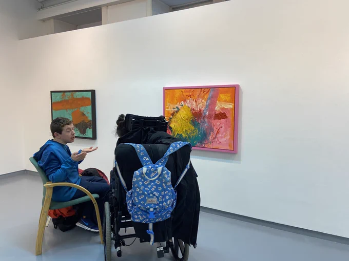 Two people sit and chat at Erin Keogh's Line, Patches and Colourhands reception, one of them on a wheelchair and the other on a chair. There are two artworks in the wall, one mainly teal and brown and the other pink and orange.