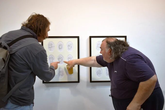 Two men point at an artwork of all of the Doctor Whos, from the Young Talent exhibition reception. One of them is wearing a backpack and the other is holding a paper cup.