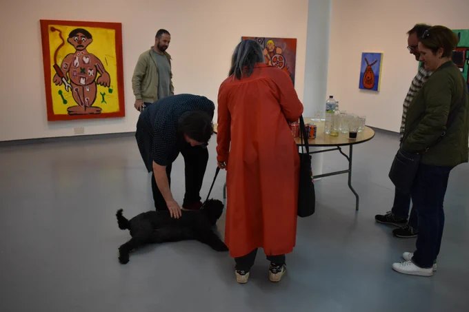 A group of people at Jonathan McKinstry's I like things I remember reception. There is a table with refreshments in the middle and Jonathan is crouching to pet a black dog.