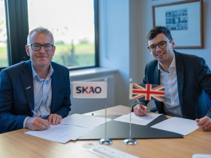 Two smiling men signing documents together at a table with two small flags depicting the SKAO and Union Jack in front of them.