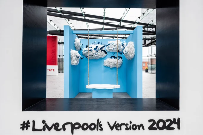 A large outdoor art installation and photo-spot. The installation looks like a giant, walk-through polaroid with a scene featuring a blue sky, clouds and birds and a swing to sit on. Black writing reads 'LIVEPROOL'S VERSION 2024.'