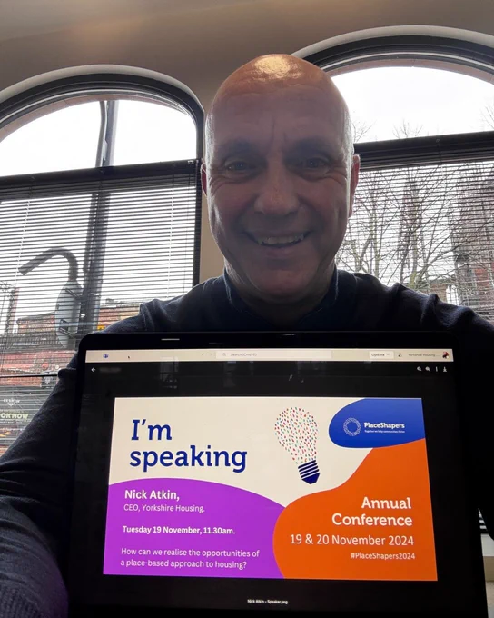 Nick Atkin sitting behind a laptop screen that's facing the camera with a multi-coloured graphic on it that reads 'I'm speaking at place shapers conference 2024'