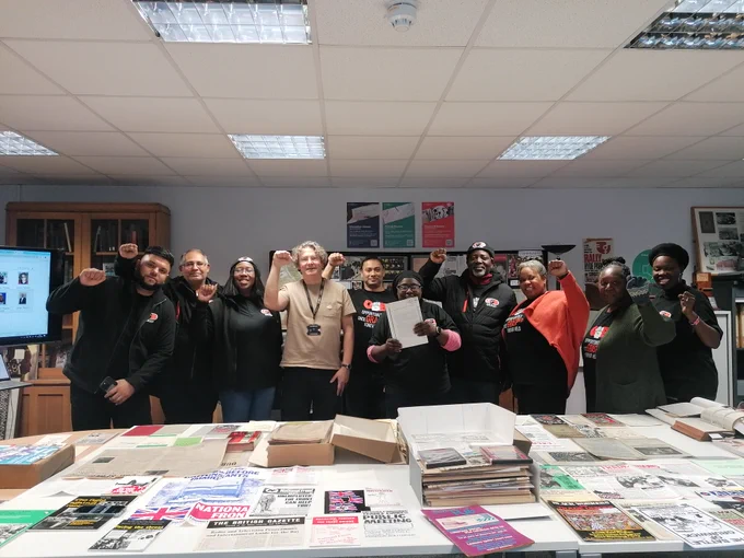 Photo showing GMB activists in the TUC Library reading room, with Jeff the librarian in the middle. All have the fists raised. The desks infront of them are filled with archive boxes
