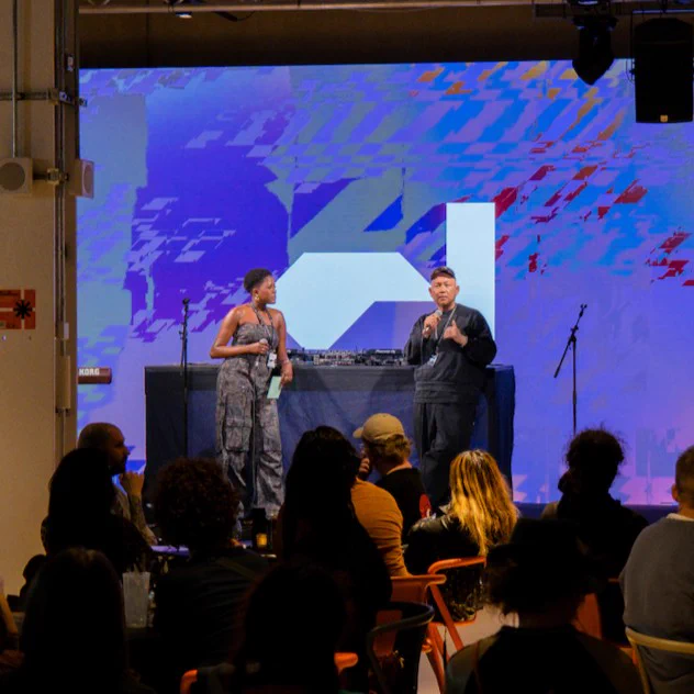 Two people speaking on stage in the Social at Aviva Studios with a a large digital screen displaying Factory International's logo in the background, and an attentive audience in the foreground.