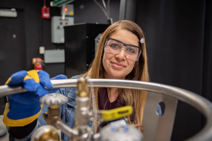 Woman with long brown hair wearing safety goggles and gloves in a lab.