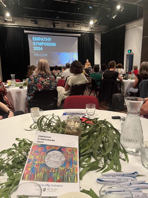 A conference room with people seated at round tables and a slide on screen saying "Empathy Symposium 2024". Gum leaves, water glasses and jugs, and an abstract book are positioned on the table.