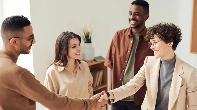 Photo of two people shaking hands while two other people stand nearby, smiling.