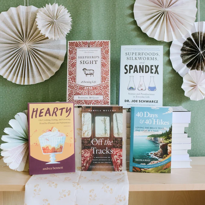 Five books are displayed in front of green fabric and surrounded by paper snowflake decorations made from book pages. The books are Shepherd's Sight by Barbara McLean, Superfoods, Silkworms, and Spandex by Dr. Joe Schwarcs, Hearty by andrea bennett, Off the Tracks by Pamela Mulloy, and 40 Days and 40 Hikes by Nicola Ross.