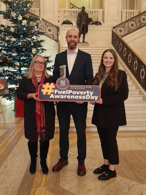 Photo of three people standing in the Great Hall in Stormont in front of the stairs and the big Christmas tree, holding a #FuelPovertyAwarenessDay sign. The people are:
Pat Austin (Director NEA NI), Mark H Durkan MLA, and Shannon Montgomery (Policy and Campaigns Officer NEA NI).