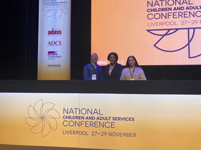 Minister for Children and Families Janet Daby MP stands in the middle of LGA chair of Children and Young People's Board, Arooj Shah & Andy Smith, President of ADASS. All three are looking to camera standing behind a large National Children and Adult Services digital conference sign.