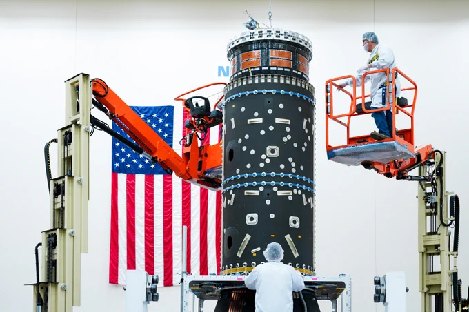 Technicians work on the installation of hardware designed to house the Power and Propulsion Element's xenon fuel tanks, an integral component of its advanced electric propulsion system. The setup is conducted in a facility, with one technician on an elevated lift and another standing at the base of the structure. An American flag is displayed prominently in the background. Credit: Maxar Space Systems