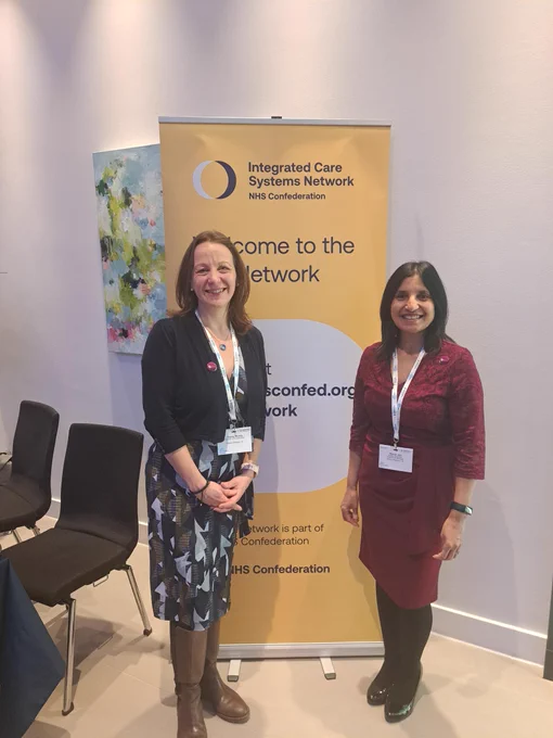 Director of programmes, Tracey, and Health equalities programme manager, Neerja, standing in front of a banner that reads "Welcome to the Integrated Care Systems Network - NHS Confederation" at a conference. They are wearing event lanyards and smiling.