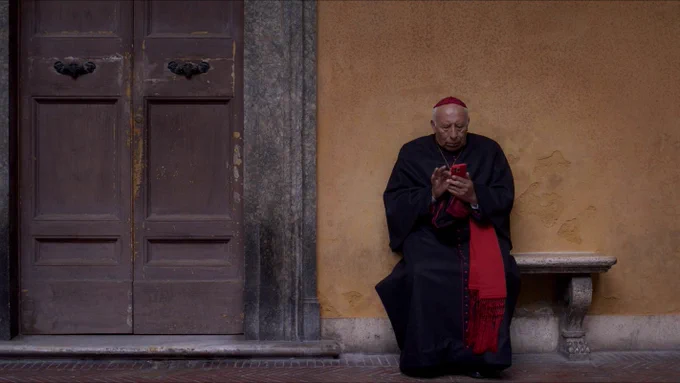 a still from the movie Conclave of a cardinal outside a building sitting looking at his phone