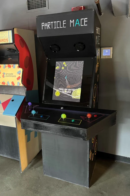 A black arcade cabinet with the words "PARTICLE MACE" on the marquee, with the second A in bright teal. You can see bright yellow shapes, on the right side, and a joystick and button set up for four players. The screen is in attract mode so there is some gameplay visible. On the left, but cut off in the picture, is another arcade cabinet called Super Sharp Birds, with a bird and egg motif in bright red and blue against white.