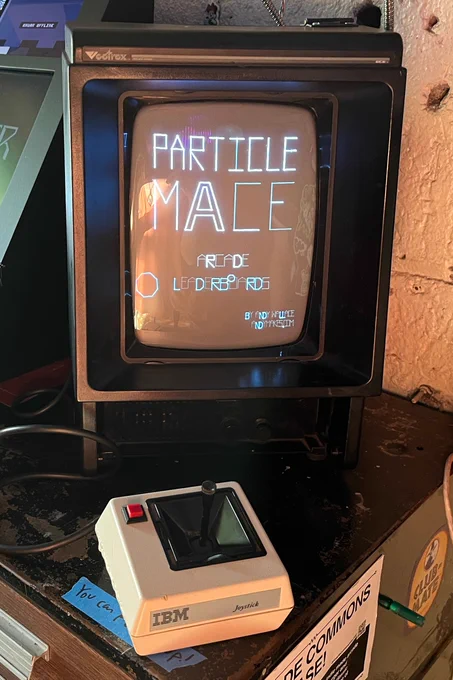 A vectrex console, which is a vector display based home video console with an in-built vertically oriented CRT monitor (so much taller than wide). It shows the words "PARTICLE MACE" on the screen. In front of it, there is an IBM joystick controller with a big red button on it.