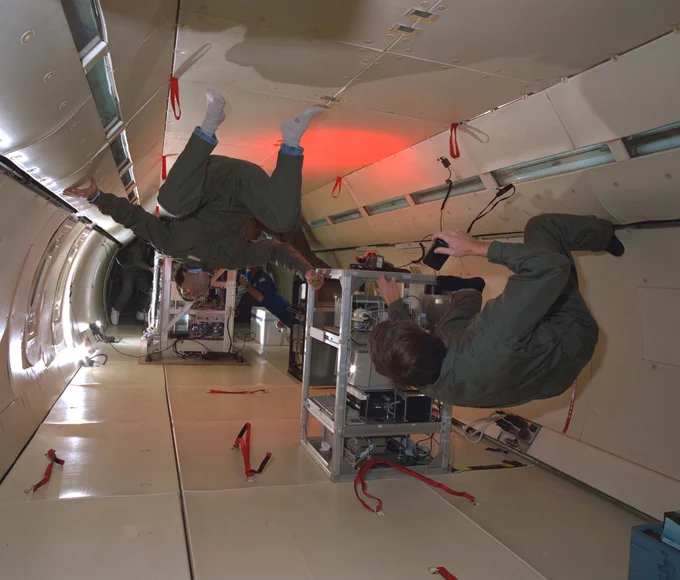 Two people wearing green flight suits float sideways and upside down inside of a plane cabin and interact with experiment equipment. Credit: NASA/Quentin Schwinn