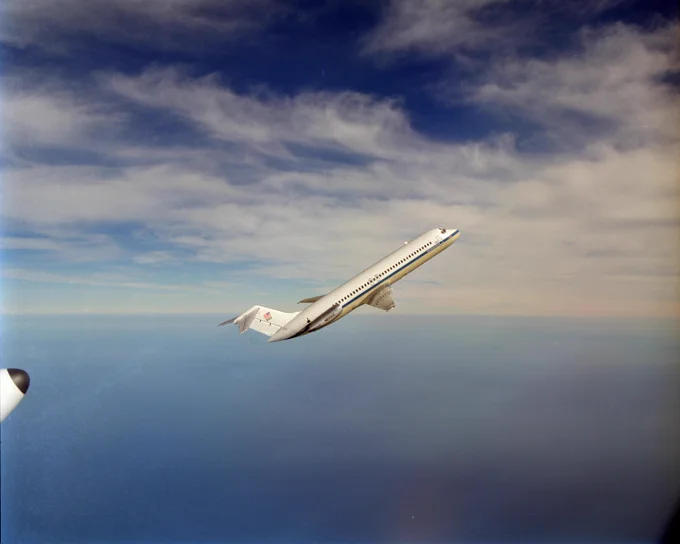 A white DC-9 plane with a blue stripe and text that says “Lewis Research Center” flies upward against a blue sky with several wispy white clouds. Credit: NASA/Quentin Schwinn