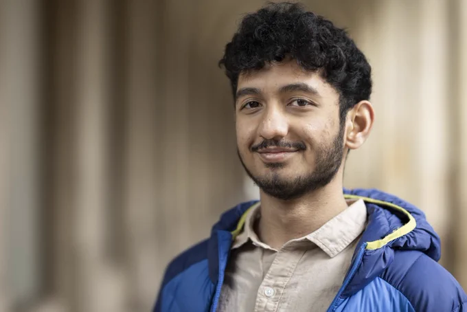 Photo of student looking directly at the camera. He's wearing a blue raincoat and a beige shirt.