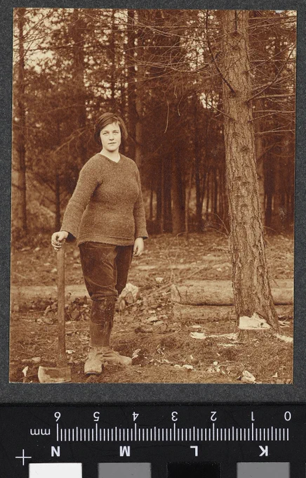 A woman dressed in breeches, knee high laced boots and jumper stands holding an axe. She is next to a tree with with the initial axe marks on it's lower trunk. The Women's Forestry Service was started by Miss Rosamund Crowdy as a section of the Land Army. It was set up under theTimber Supply Department, part of the Board of trade in 1917. Training camps for cutting and measuring the timber were held at several locations, including Wendover.