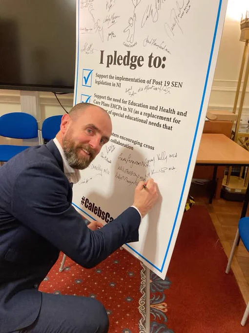 Mark H Durkan MLA kneeling in front of and signing a pledge board, reaffirming his support for the Caleb's Cause Campaign.
