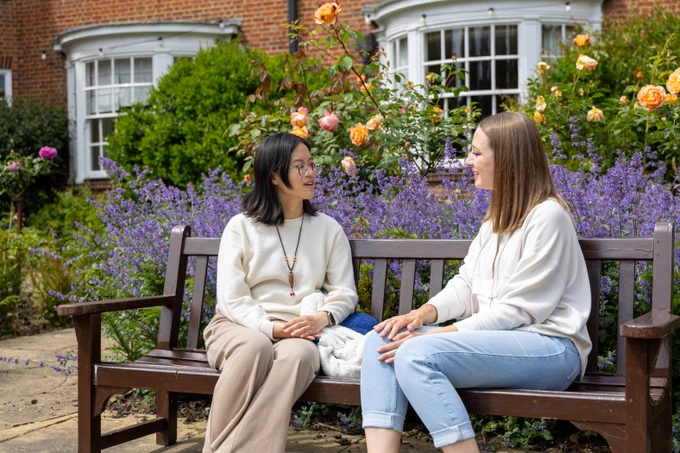 Two people sitting on a bench, conversing in a garden with blooming roses and purple flowers, with a brick building in the background.