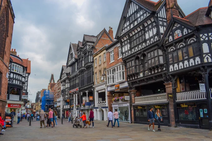 A bustling high street with people walking and shopping, lined with historic black-and-white Tudor-style buildings.