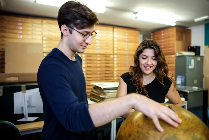 Two students in the Department's Map Library engaging with a large vintage globe. The background features wooden drawers and desks, One student points at the globe while the other smiles.