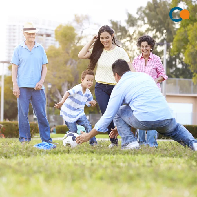 Family playing football together in the park on a sunny day; features four adults, one child. 