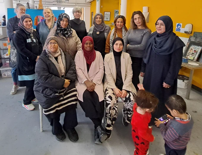 A group of women sit and stand posing for a photo in an arts studio. There are two kids in the forefront, both looking at a game in a phone.